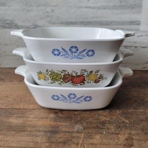 May include: A stack of three white ceramic baking dishes with integrated side handles. The top and bottom dishes feature a blue floral pattern, while the middle dish displays a colorful botanical illustration of various vegetables and fruits.