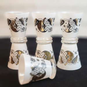 May include: Set of four white shot glasses with gold and black abstract designs. Each glass features a stylized fruit with leaves and the word "ginger" written on it.
