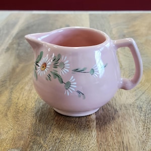 May include: A pink ceramic creamer with a floral design. The creamer has a handle and a rounded body. The flowers are white with green leaves.