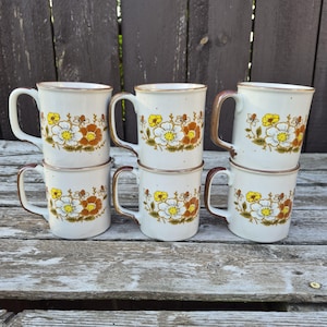 May include: A set of six off-white ceramic mugs arranged in two rows. Each mug features a repeating floral pattern with yellow, white, and brown flowers and green leaves. The mugs have a brown rim and a matching brown handle on the side.