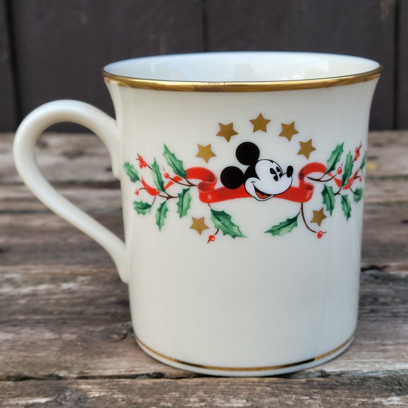 May include: White ceramic mug with a gold rim and a Christmas-themed design featuring Mickey Mouse wearing a red ribbon and surrounded by holly and gold stars.
