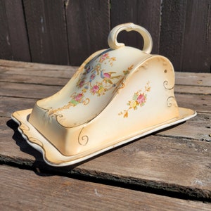 May include: A vintage ceramic butter dish with a floral design. The dish has a domed lid with a handle and a base with a scalloped edge.