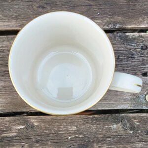 May include: A white ceramic mug with a gold rim. The mug is empty and has a slight reflection in the bottom.