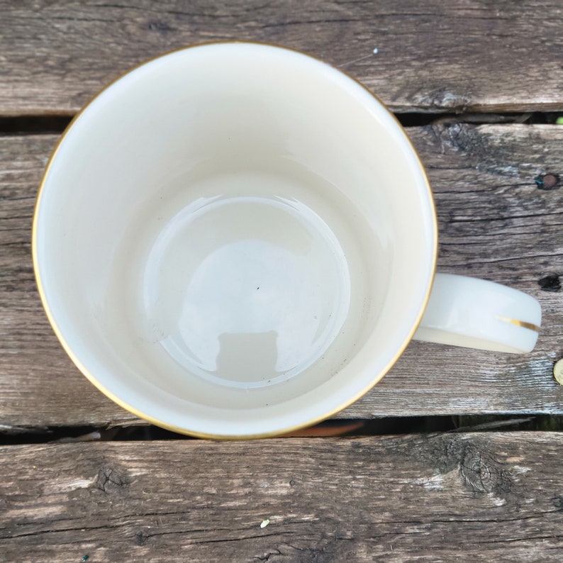 May include: A white ceramic mug with a gold rim. The mug is empty and has a slight reflection in the bottom.