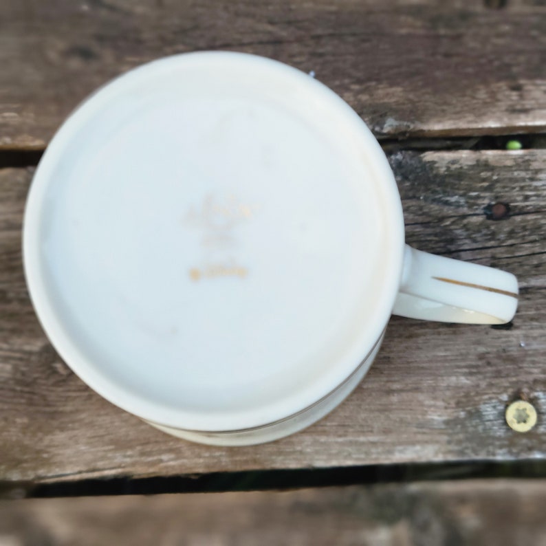 May include: White ceramic teacup with a gold rim and a gold inscription on the bottom. The teacup has a handle.