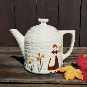 May include: White ceramic teapot with a brick pattern and a woman holding flowers. The teapot has a brown handle and a brown spout. The teapot is sitting on a wooden surface.