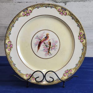 May include: A vintage decorative plate with a yellow center and a detailed border. The border features pink floral designs and gold accents. The center of the plate showcases two birds perched on a branch with pink blossoms.