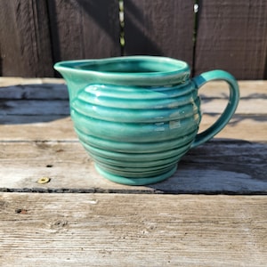 May include: A turquoise ceramic creamer with a handle and a spout. The creamer has a ribbed design around the body. The creamer is sitting on a weathered wooden surface.