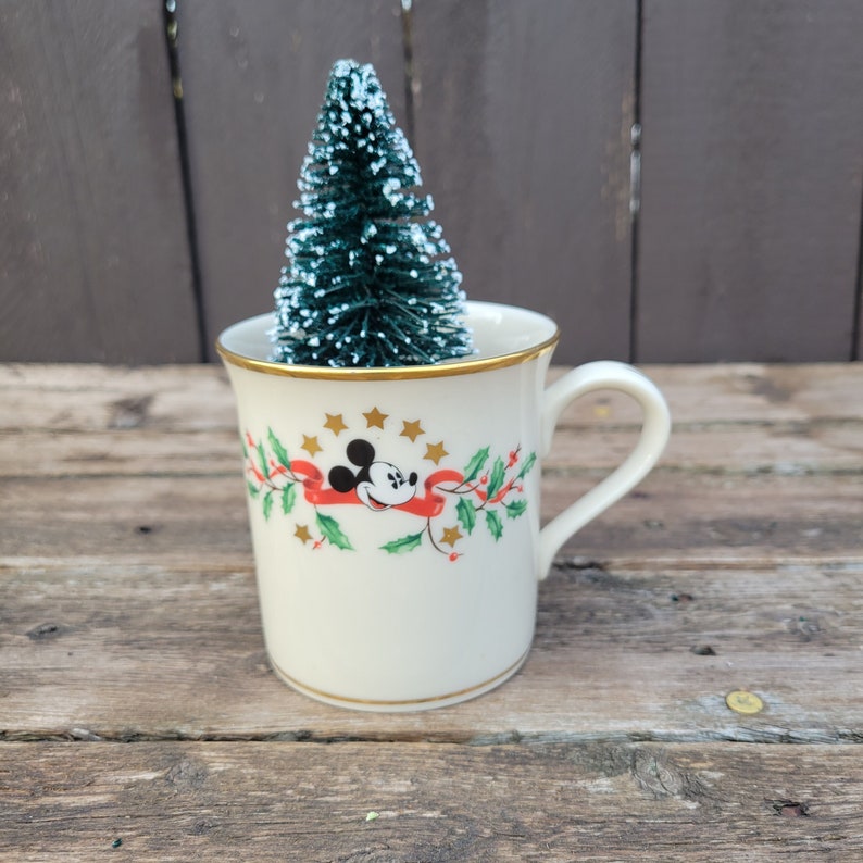 May include: A white ceramic mug with a gold rim and a Christmas design featuring Mickey Mouse, holly, and a red ribbon. A small green artificial Christmas tree is inside the mug.
