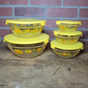 May include: A set of clear glass mixing bowls with yellow lids. Each bowl is decorated with a sunflower design. The bowls are in various sizes, stacked on a wooden surface, with a brick background.