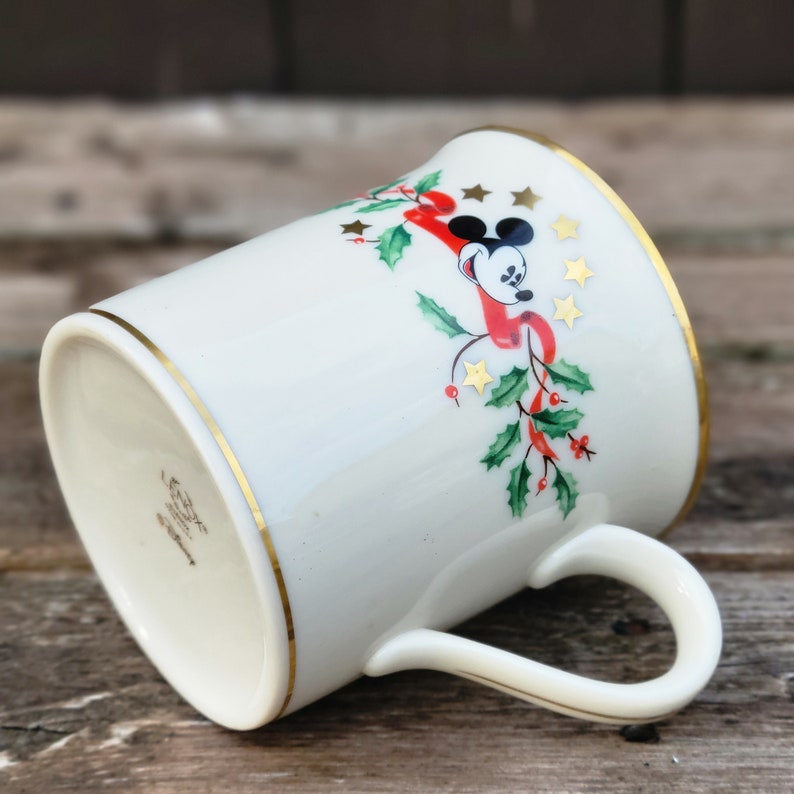 May include: White ceramic mug with a gold rim and a Christmas-themed design featuring Mickey Mouse, holly, and red ribbon. The mug has a gold band around the bottom and a gold handle.