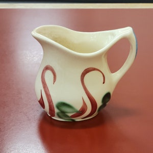 May include: A small white ceramic creamer with a red and green floral design. The creamer has a handle and a rounded body.