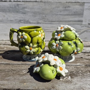 May include: A ceramic tea set featuring a mug, a sugar bowl with a lid, and a small creamer. The set is a vibrant green with white daisy accents and orange centers. The pieces are arranged on a wooden surface.