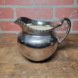 May include: A silver-plated creamer with a rounded body, a spout, and a curved handle. The rim of the creamer has a decorative beaded edge. The creamer is filled with a light-colored liquid. The background is a brick wall and a wooden surface.