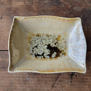 May include: A rectangular ceramic dish with a cream-colored base and a textured, crystalline design in the center. The dish has a glossy finish with brown and gold accents along the edges. The dish is on a wooden surface.