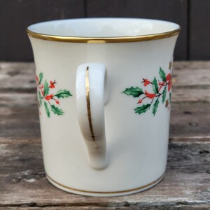 May include: White ceramic mug with a gold rim and handle. The mug has a Christmas holly design with red berries and green leaves.