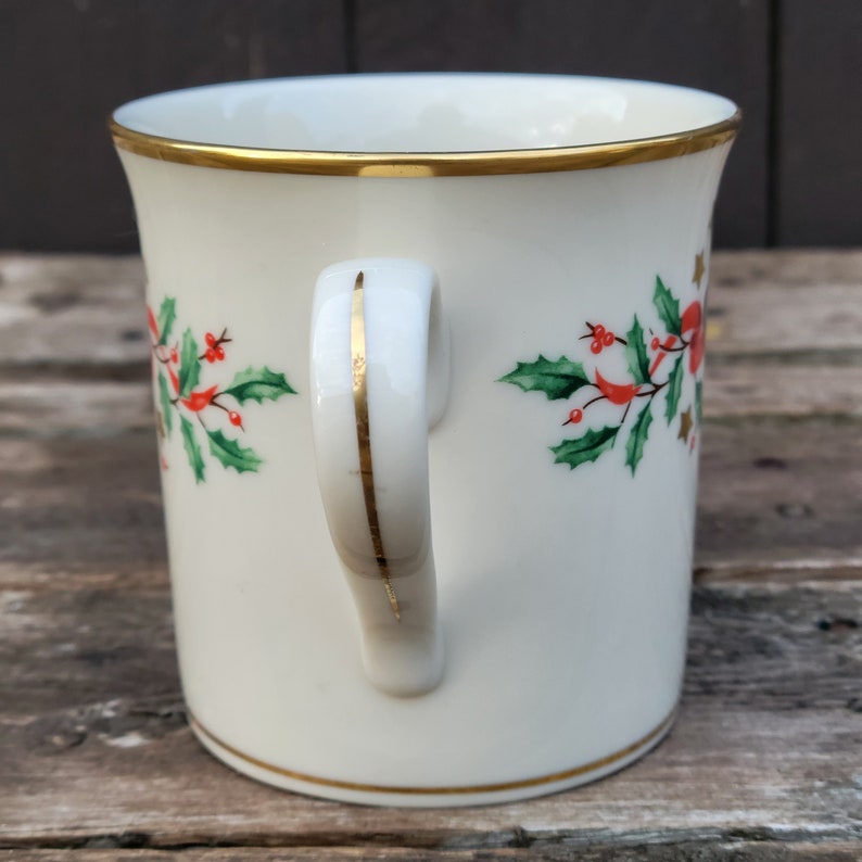 May include: White ceramic mug with a gold rim and handle. The mug has a Christmas holly design with red berries and green leaves.