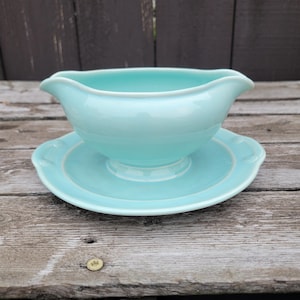 May include: A light blue ceramic gravy boat with a matching saucer. The gravy boat has a curved spout and a pedestal base. The saucer has a scalloped edge. The set is displayed on a weathered wooden surface.