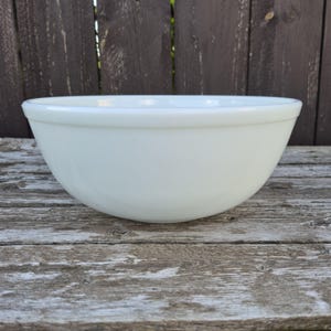 May include: A solid white mixing bowl with a smooth, glossy finish and a rounded, deep basin. The bowl features a simple, clean design with a slightly flared rim, providing a wide opening for mixing ingredients while maintaining a sturdy structure.