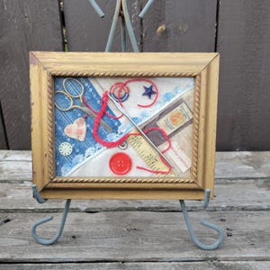 May include: A framed collage featuring sewing notions. The gold-toned frame surrounds a display of scissors, a crochet hook, red thread, a measuring tape, buttons, and fabric pieces in blue and white patterns. A small metal easel supports the frame.