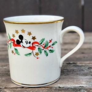 May include: A white ceramic mug with a gold rim and handle. The mug features a festive design with Mickey Mouse, holly, and red ribbon. There are gold stars scattered around the design.