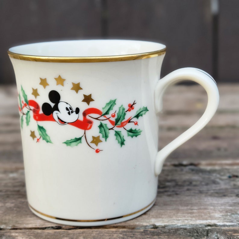 May include: A white ceramic mug with a gold rim and handle. The mug features a festive design with Mickey Mouse, holly, and red ribbon. There are gold stars scattered around the design.