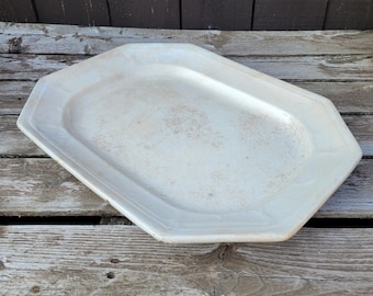 May include: A large, off-white, octagonal serving platter. The platter has a slightly raised rim and a smooth, slightly stained surface. The shape is geometric, and the overall aesthetic is vintage.