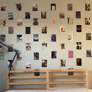 May include: A wooden bed frame with a white mattress and a wall covered in framed photos. The photos are of various sizes and subjects, including cityscapes, landscapes, and abstract art. The wall is painted a light gray color. The bed frame has a simple design with four horizontal slats.