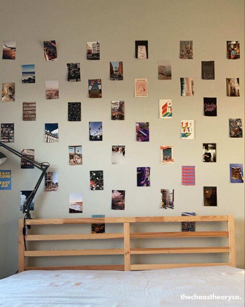 May include: A wooden bed frame with a white mattress and a wall covered in framed photos. The photos are of various sizes and subjects, including cityscapes, landscapes, and abstract art. The wall is painted a light gray color. The bed frame has a simple design with four horizontal slats.