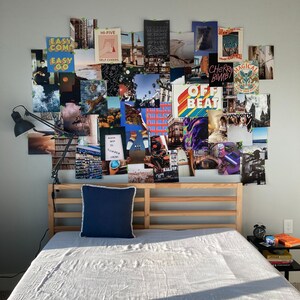 May include: A wooden bed frame with a white linen duvet and a blue pillow. The wall behind the bed is covered in a collage of colorful posters with various text and images, including "Easy Come Easy Go", "Hi-Five", "Self Cheers", "Cherry Bomb", "Magical", "Off Beat", and "Weekend".