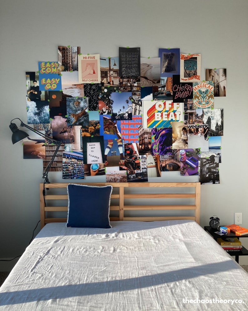 May include: A wooden bed frame with a white linen duvet and a blue pillow. The wall behind the bed is covered in a collage of colorful posters with various text and images, including "Easy Come Easy Go", "Hi-Five", "Self Cheers", "Cherry Bomb", "Magical", "Off Beat", and "Weekend".