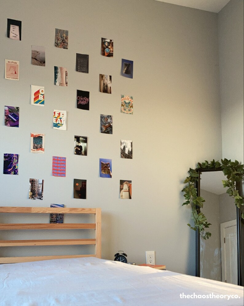 May include: A white bed with a wooden headboard and a white bedspread. The wall behind the bed is decorated with a variety of framed photos and prints. A large mirror with a leafy vine is in the corner of the room.