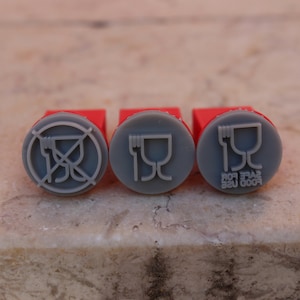 May include: Three red rubber stamps with grey circular designs. The first stamp says "Food Safe For Use". The second stamp has a picture of a fork and a cup. The third stamp has a picture of a fork and a cup with a diagonal line through it.