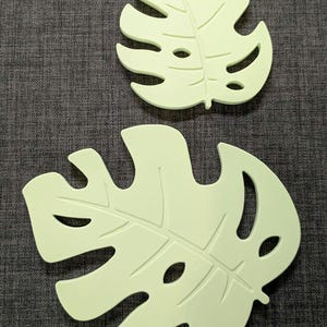 Peut inclure: Deux dessous de plat en forme de feuilles vert clair. Les dessous de plat sont conçus pour ressembler à des feuilles de Monstera, avec des sections découpées et des motifs de veines détaillés. Ils sont en matière souple, posés sur une surface grise texturée.