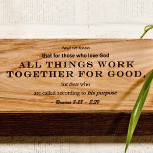 Puede incluir: Un letrero rectangular de madera con el texto "ALL THINGS WORK TOGETHER FOR GOOD" en letras negras. El letrero también incluye el versículo bíblico Romanos 8:28. El letrero se muestra en un estante de madera oscura.