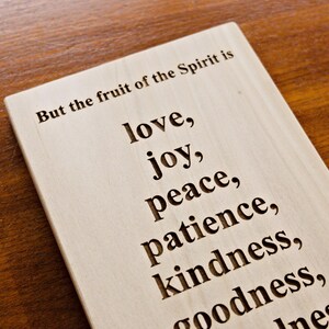 May include: Wooden plaque with engraved text. The text reads "But the fruit of the Spirit is love, joy, peace, patience, kindness, goodness..." The plaque is light-colored and set against a dark wood surface.