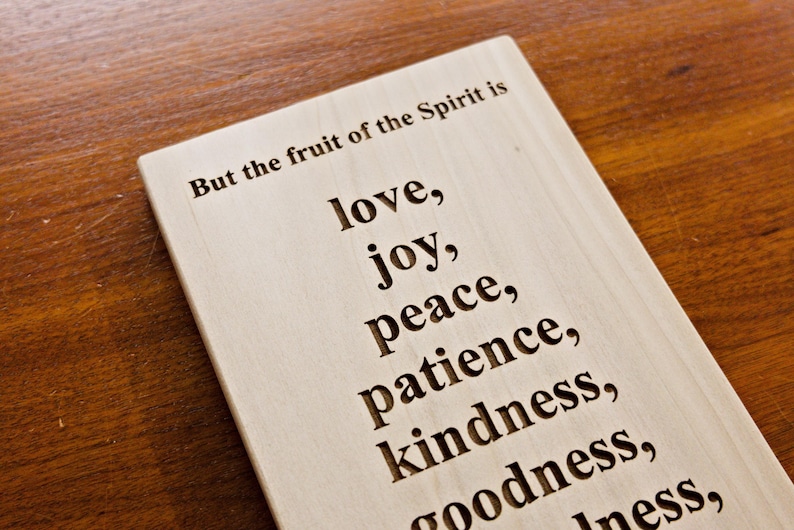 May include: Wooden plaque with engraved text. The text reads "But the fruit of the Spirit is love, joy, peace, patience, kindness, goodness..." The plaque is light-colored and set against a dark wood surface.