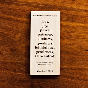 May include: A rectangular wooden sign with the text "But the fruit of the Spirit is love, joy, peace, patience, kindness, goodness, faithfulness, gentleness, self-control; against such things there is no law. Galatians 5:22-23". The sign is on a wooden surface.