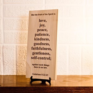 May include: A wooden plaque with the text "But the fruit of the Spirit is love, joy, peace, patience, kindness, goodness, faithfulness, gentleness, self-control; against such things there is no law. Galatians 5:22-23". The plaque is displayed on a black stand.