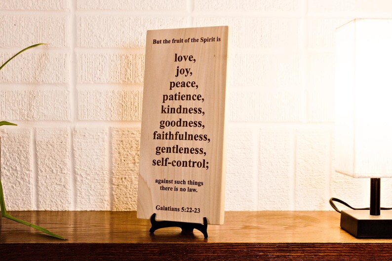May include: A wooden plaque with the text "But the fruit of the Spirit is love, joy, peace, patience, kindness, goodness, faithfulness, gentleness, self-control; against such things there is no law. Galatians 5:22-23". The plaque is displayed on a black stand.