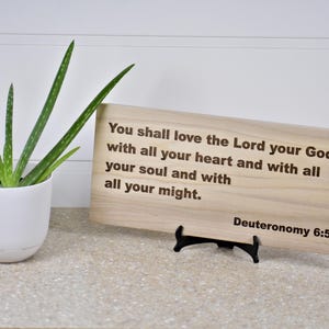 Puede incluir: Una placa de madera con una cita religiosa grabada. El texto dice: «Amarás al Señor tu Dios... Deuteronomio 6:5». A la izquierda, una planta de aloe vera en maceta.
