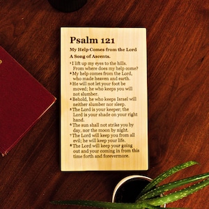 Puede incluir: Una placa de madera con el texto del Salmo 121, un himnario rojo y una pequeña planta en maceta sobre una superficie de madera oscura. La placa presenta el título y los versículos del pasaje bíblico.