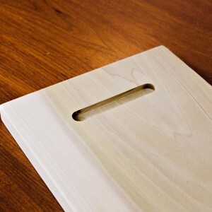 May include: A light-colored wooden plaque with a rectangular shape and a routed slot for hanging. The wood grain is visible, and the plaque is set against a dark wood surface.