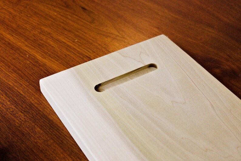 May include: A light-colored wooden plaque with a rectangular shape and a routed slot for hanging. The wood grain is visible, and the plaque is set against a dark wood surface.