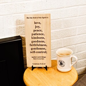 May include: A wooden sign with the text "But the fruit of the Spirit is love, joy, peace, patience, kindness, goodness, faithfulness, gentleness, self-control;" and a white mug with a logo on a wooden table.