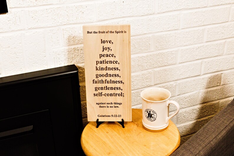 May include: A wooden sign with the text "But the fruit of the Spirit is love, joy, peace, patience, kindness, goodness, faithfulness, gentleness, self-control;" and a white mug with a logo on a wooden table.