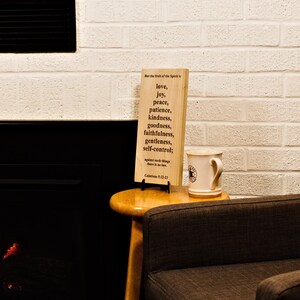 May include: A wooden sign with the words "love, joy, peace, patience, kindness, goodness, faithfulness, gentleness, self-control" printed on it. A white mug with a black logo sits next to the sign on a small wooden table. The background is a white brick wall.