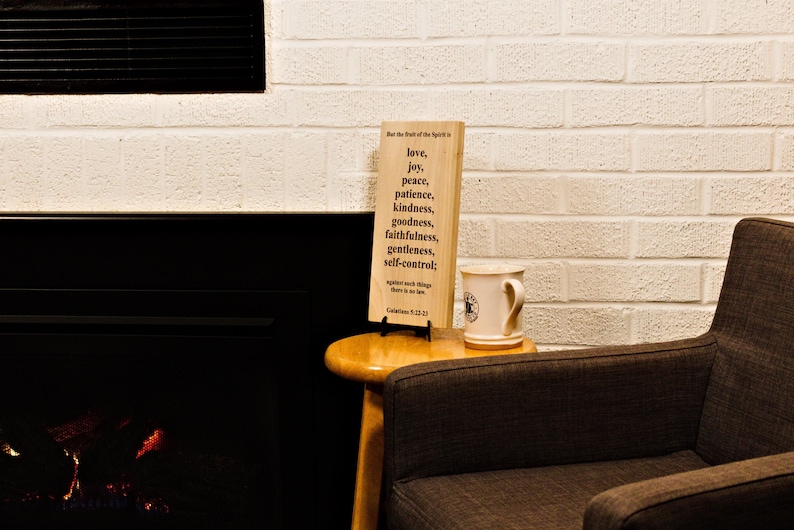 May include: A wooden sign with the words "love, joy, peace, patience, kindness, goodness, faithfulness, gentleness, self-control" printed on it. A white mug with a black logo sits next to the sign on a small wooden table. The background is a white brick wall.