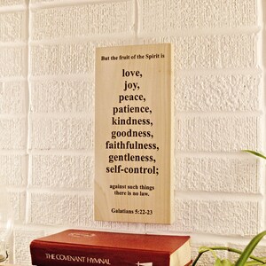 May include: A wooden sign with the text "But the fruit of the Spirit is love, joy, peace, patience, kindness, goodness, faithfulness, gentleness, self-control;" against a white brick wall. A red book is at the bottom.