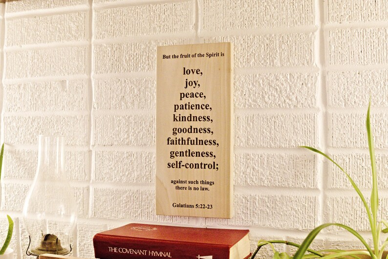 May include: A wooden sign with the text "But the fruit of the Spirit is love, joy, peace, patience, kindness, goodness, faithfulness, gentleness, self-control;" against a white brick wall. A red book is at the bottom.